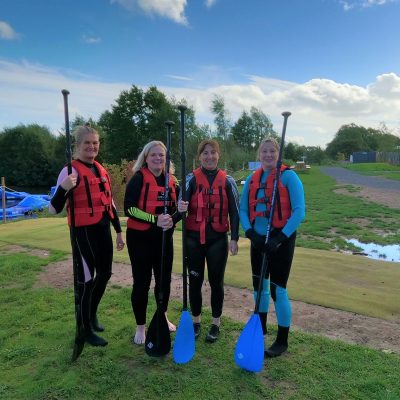 Four women wearing life jackets and holding paddles on a grassy area near water.