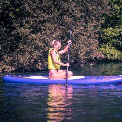 A woman in a yellow life vest paddleboards on calm water.