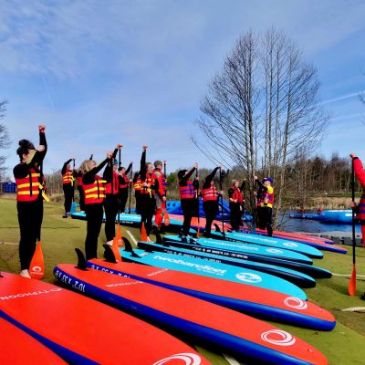 A group of people in life jackets and wetsuits stand with their paddleboards, raising their paddles.