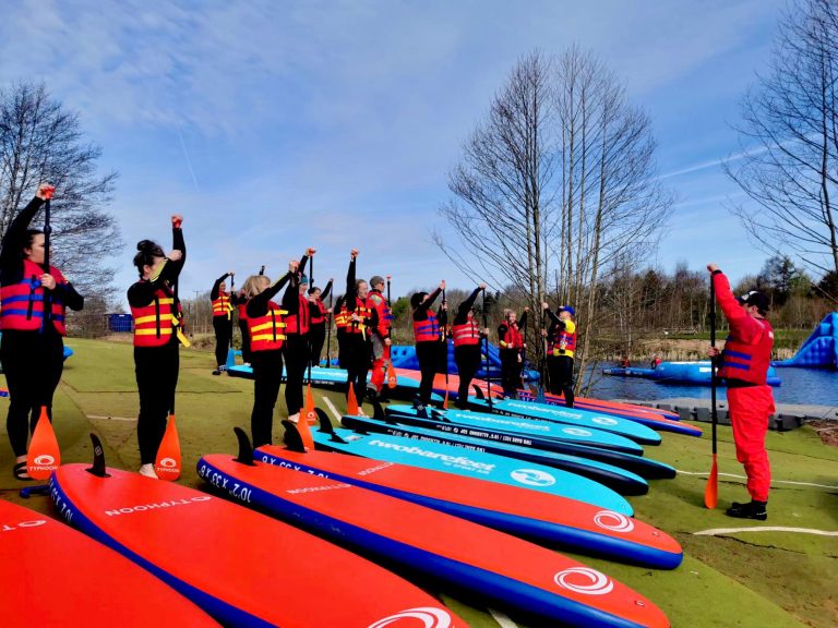 A group of people in life jackets and wetsuits stand with their paddleboards, raising their paddles.