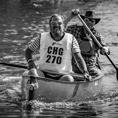 A man paddles a canoe while another sits in the back, both wearing life vests.