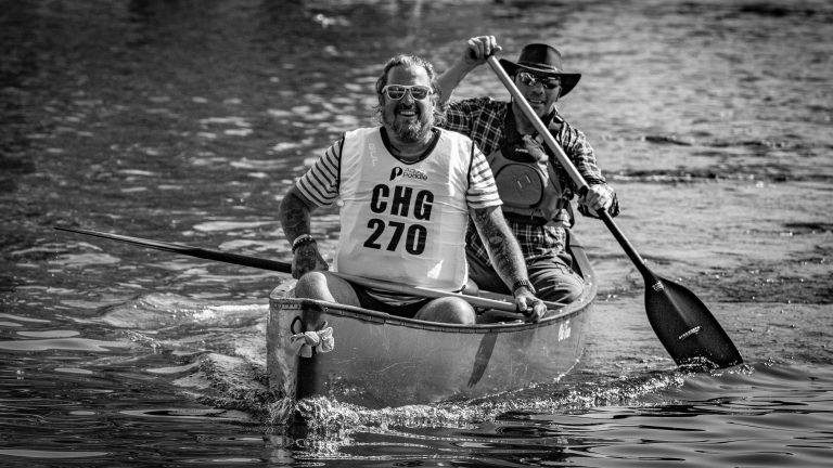 A man paddles a canoe while another sits in the back, both wearing life vests.