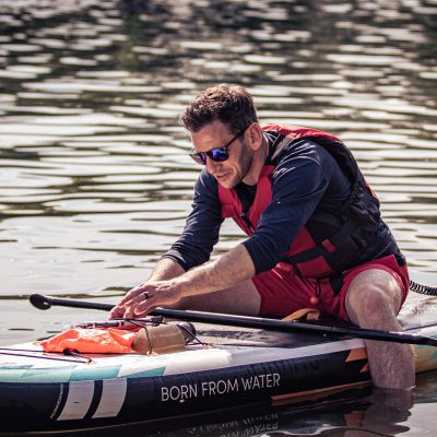 A man in a life jacket and sunglasses sits on a stand-up paddleboard, adjusting his gear.