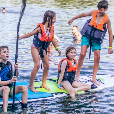Four children wearing life jackets and standing or sitting on stand-up paddleboards in a body of water.