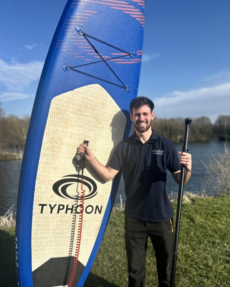 A smiling man holds a paddle and stands next to a blue Typhoon brand stand-up paddleboard.