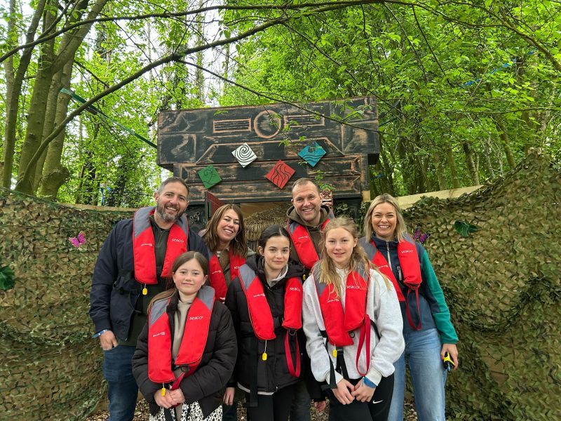 A group of people wearing life jackets pose in front of a wooden sign with various decorations.