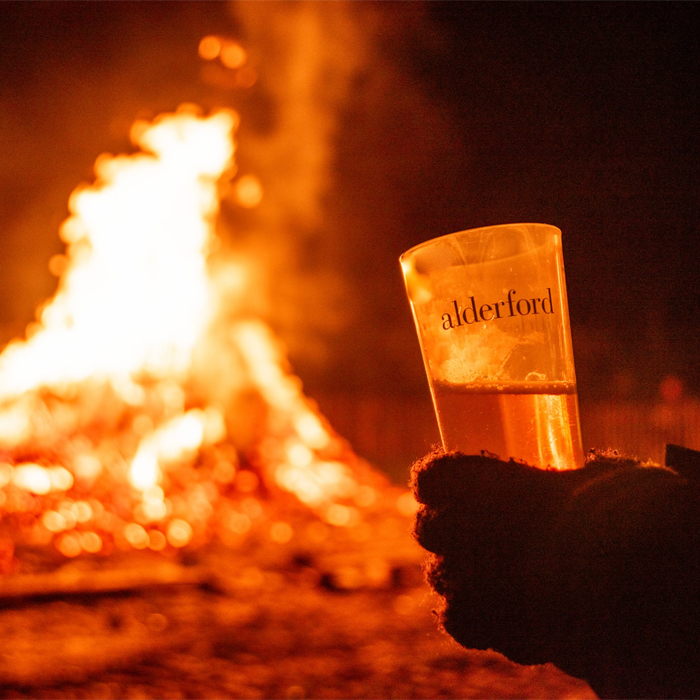 a hand holding a glass of Alderford beer in front of a bonfire with flames visible behind it.