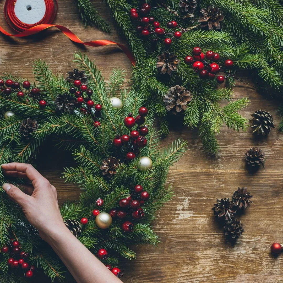 A hand is seen adjusting a red and green holiday wreath with pine cones, berries, and ornaments on a wooden surface.
