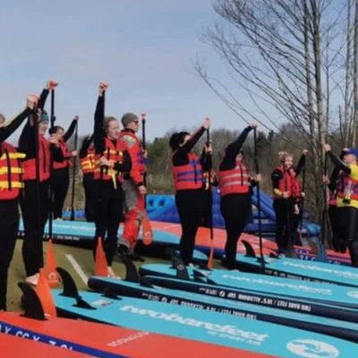 A group of people in life jackets and wetsuits stand on paddleboards, raising their paddles.
