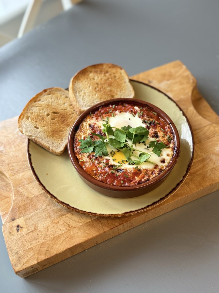 A bowl of Eggs Benedict garnished with herbs and a slice of butter, served on a plate next to two slices of toast.