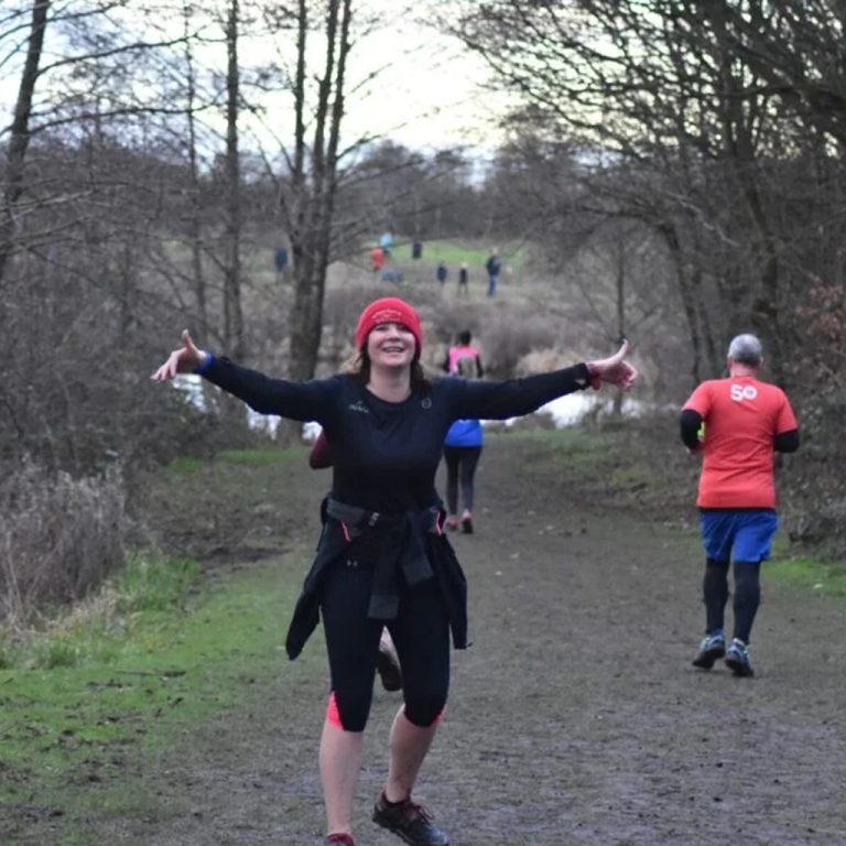 A woman wearing a red beanie, black long-sleeve top and leggings is standing on a trail with her arms wide open.