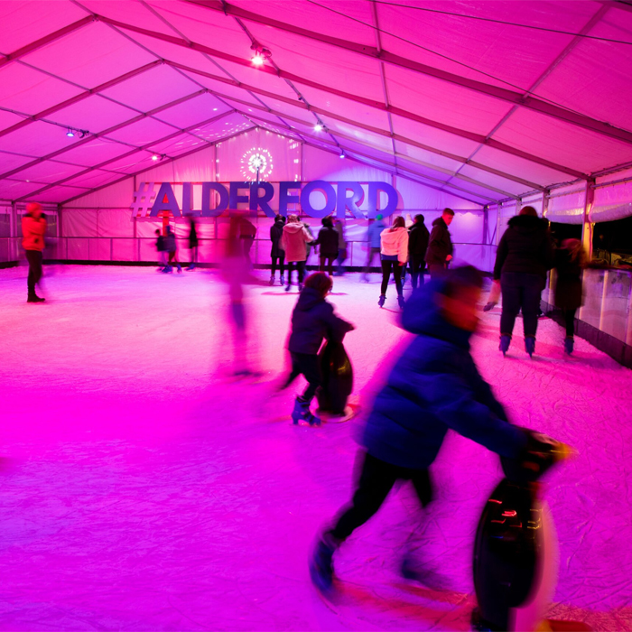People ice skating in a pink-lit tent with 'Alderford' sign.