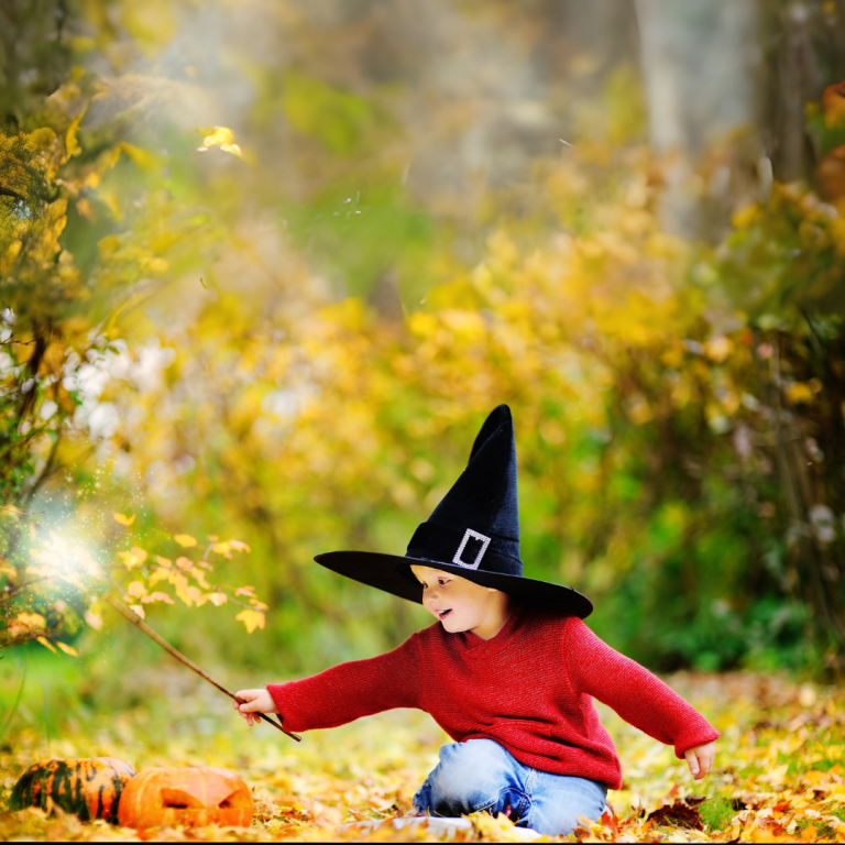 A child wearing a witch hat and red sweater is playing with a sparkler on the ground, surrounded by autumn leaves.