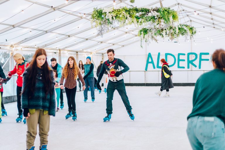 A group of people ice skating on a rink with 'ALDERF' written in the background.