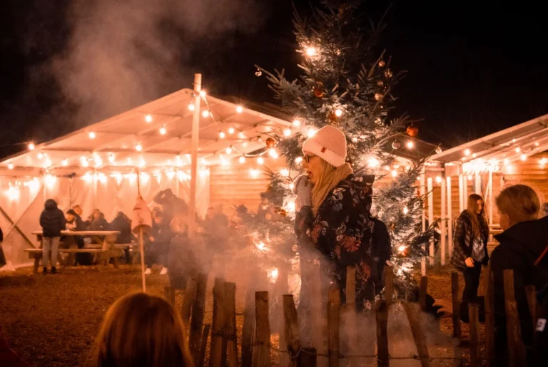 A woman in a white hat and floral coat stands near a lit-up Christmas tree at an outdoor market.