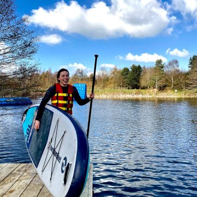 A woman in a life jacket holding a paddle and standing on a dock with a stand-up paddleboard.