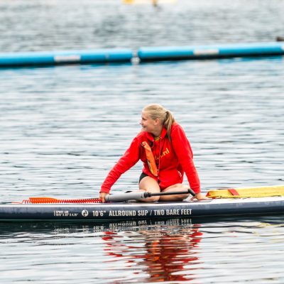 A woman in a red jacket sits on a paddleboard with blue water and floating lane markers behind her.
