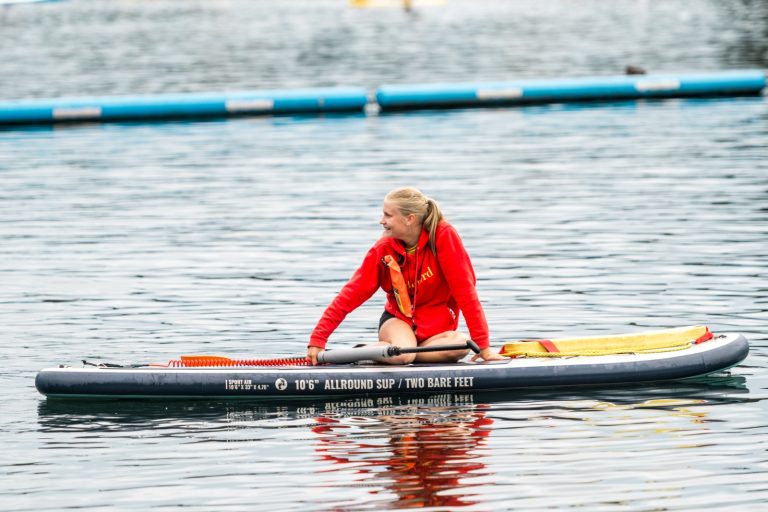 A woman in a red jacket sits on a paddleboard with blue water and floating lane markers behind her.